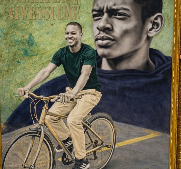 Okeke Kosisochukwu Victory's framed artwork: a young Black cyclist in a green shirt and pale trousers, riding a bicycle, with a second close-up portrait of his determined face superimposed in the background. Caption: Impossible is...
