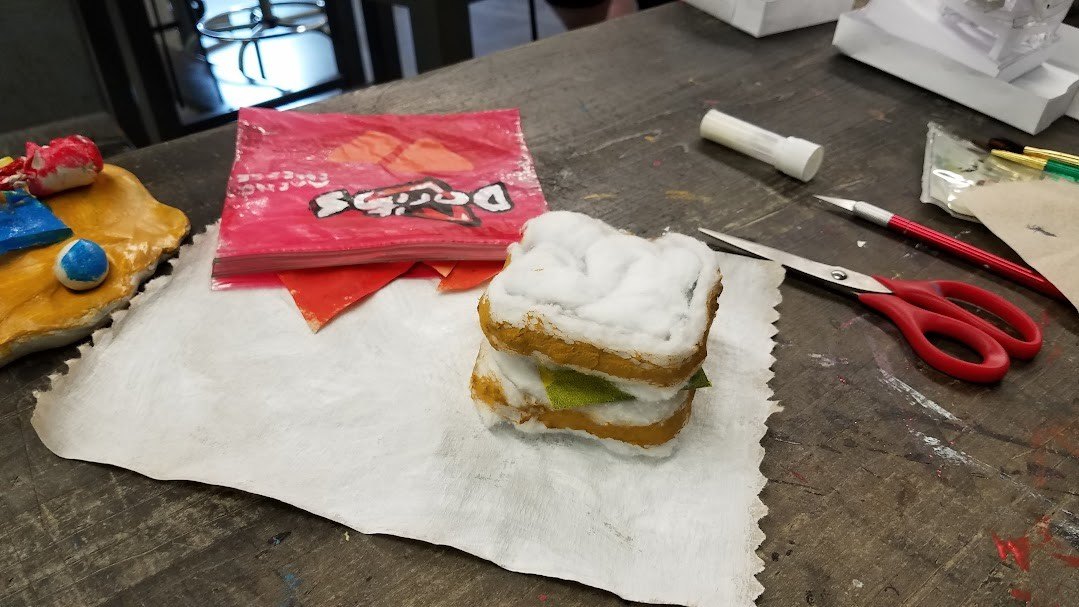 A sculpture of a packed lunch made from paper and cotton: a sandwich and a bag of chips on a napkin.