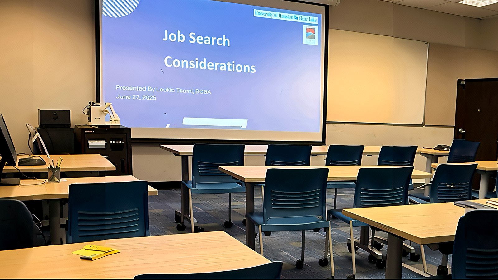 A university classroom with wooden tables and blue chairs. The projector screen at the front shows a title slide reading Job Search Considerations, presented by Loukia Tsami, BCBA, June 27, 2025.